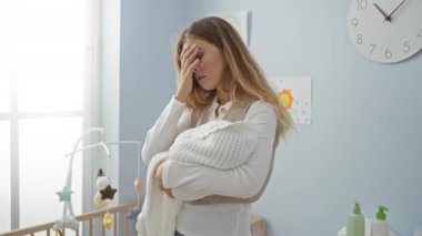 Parlak bir kreşte bebeği tutan kadın beşikte ve oyuncakların yanında bitap görünüyor. Elleri alnında duvardaki saatin altında, arka planda sanat eserleri var..