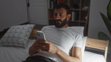 Yatak odasında akıllı telefon kullanan sakallı genç adam, loş bir odada hayal kırıklığı ve stres duygularını göstererek yatakta yatıyor.