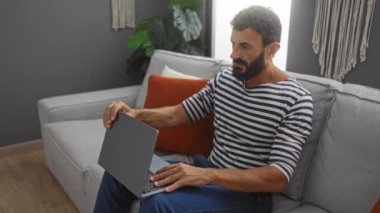 Sakallı genç adam, günlük kıyafetlerle kanepede oturuyor, bilgisayarını kapatıyor ve esniyor, bir apartman dairesinde dinleniyor..