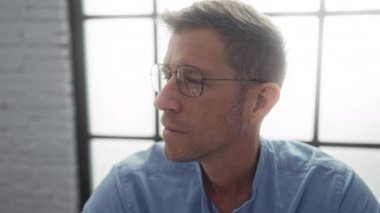 Ofiste gözlük takan genç bir adam, odaklanmış ve düşünceli, modern bir iç mimariye sahip, çekici özelliklerini ve profesyonel davranışlarını vurguluyor..