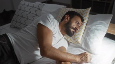 Yatak odasında sakallı genç bir adam. Hafif ışıkla aydınlatılmış. Yatağın başucundaki bir şeye uzanıyor gibi görünüyor..