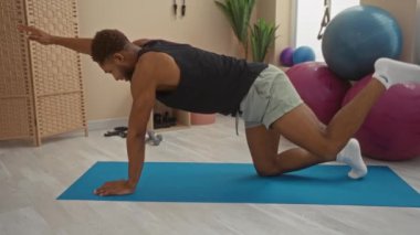 Spor salonundaki paspasın üzerinde egzersiz topları ve arka planda spor aletleri ile egzersiz yapan genç bir adam.