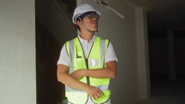 Miğfer ve güvenlik yeleği giyen genç Çinli adam güvenle kollarını kavuşturarak kapalı bir inşaat alanında duruyor.