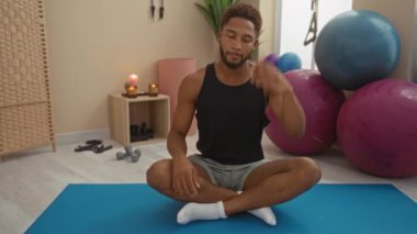 Spor salonunda arka planda egzersiz topları ve mumlarla esneyen genç bir adam mavi bir paspasla boyun egzersizi yapıyor.