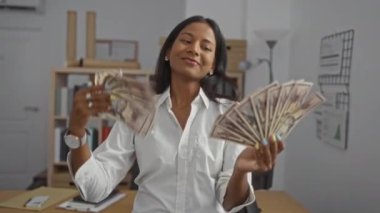 Ofiste Amerikan Doları banknotları tutan genç bir kadın, kapalı alanda profesyonel bir ortamda merak ve neşe ifadelerini yakalıyor.