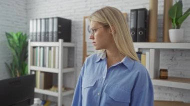 Modern ofis ortamında duran, arka planda rafları ve çiçekleri olan, gözlüklü, mavi gömlekli, odaklanmış ve profesyonel görünen bir kadın..