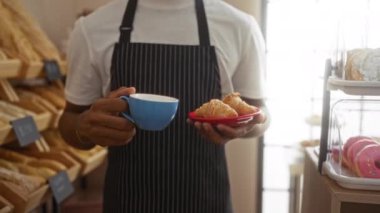 Bir fırında kahve ve kruvasan tutan, arka planda ekmek rafları olan çizgili önlük giyen ve kapalı bir kafede gülümseyen genç bir adam.