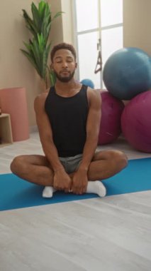 Spor salonundaki genç adam yoga minderi üzerinde meditasyon yapıyor. Egzersiz topları ve arka planda kapalı alanda bitki yetiştiriyor.