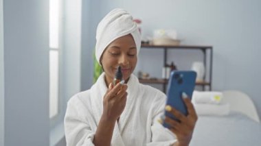 Kafasında havluyla spa odasında elinde deri şişesiyle telefona gülümseyen bir kadın.