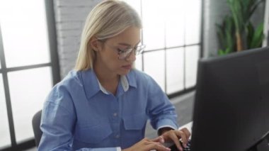 Gözlüklü, sarı saçlı, parlak, modern ofis ortamında bilgisayar başında çalışan, konsantrasyon ve profesyonelliği ön plana çıkaran bir kadın..