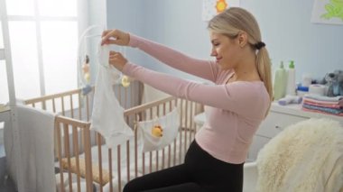 Hamile kadın rahat bir yatak odasında tahta bir beşikte oturuyor, bebek kıyafetleri tutuyor, yumuşak oyuncaklar ve bebek malzemeleriyle çevrili, sevgi dolu bir ortam yaratıyor..