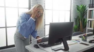 Modern ofisteki belgeleri inceleyen kadın, bilgisayarın yanında gözlük ve mavi gömlekli, geniş pencereli, çalışma ortamını ve profesyonel odaklanmayı vurgulayan doğal ışık altında..