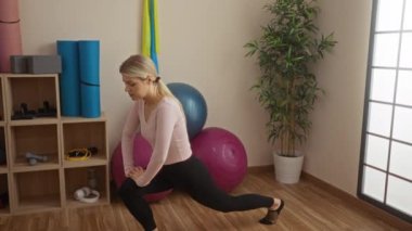 Spor salonunda pembe bluz giyen, yoga minderleri ve spor toplarıyla kendini modern, ferah bir iç mekâna saran bir kadın..