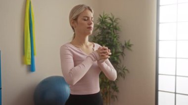 Spor salonunda meditasyon yapan bir kadın, spor aletleri ve arka planda bitkilerle rahatlama dengesi ve fitness hedefleri sergiliyor.