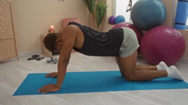 Spor salonundaki mavi minderde yoga yapan genç bir adam. Arka planda spor topları ve bitkiler var.