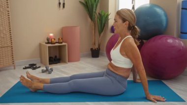 Spor salonundaki yoga minderinde egzersiz yapan olgun bir kadın. Egzersiz topları, mumlar ve spor aletleriyle.