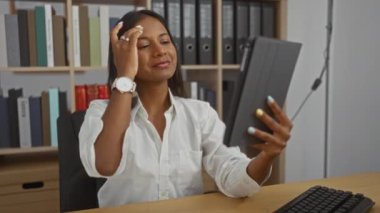 Ofis ortamında tabletle selfie çeken, gülümseyen ve saçları ayarlayan, kitaplıklar ve iş yeriyle çevrili, özgüven ve profesyonellik sergileyen bir kadın..