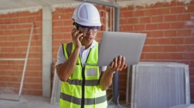 İç inşaat alanında dizüstü bilgisayar ve telefon kullanan genç bir adam.