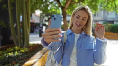 Şehir parkında akıllı telefonuyla selfie çeken bir kadın açık hava ortamında mavi gömlek giyiyor, şehir atmosferinde güneş ışığı ve yeşil ağaçlarla neşeli bir anı yakalıyor..