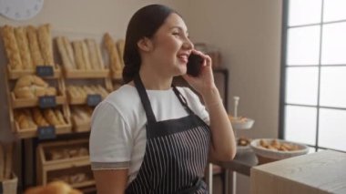 Pastanenin iç kısımlarında konuşan bir kadın ekmek raflarıyla çevrili önlük giyiyor.