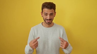 Şakacı bir ifadeye sahip genç adam kendini işaret ediyor ve parmaklarıyla hareketler yapıyor, canlı sarı bir arka plana gülümsüyor, neşeli ve komik bir an yaratıyor..