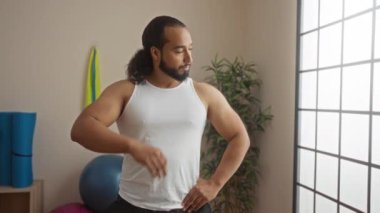 Genç adam alnındaki teri siliyor, sağlık merkezinde duruyor, beyaz bir atlet giyiyor ve arka planda egzersiz ekipmanları ve bitkiler var..