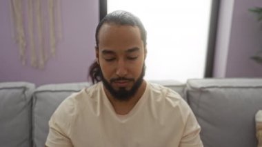 Oturma odasında rahat koltukta oturan sakallı genç adam kameraya gülümsüyor, mor duvarlar ve doğal aydınlatma sıcak, davetkar bir atmosfer yaratıyor..