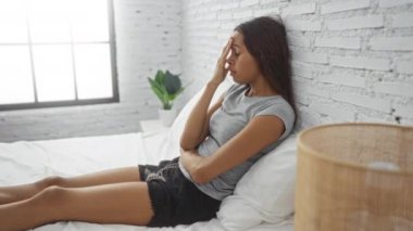 Yatak odasında, gri duvarları olan bir yatakta oturan genç bir kadın derin düşüncelere dalmış.