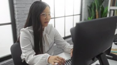 Modern bir ofiste çalışan bir kadın, büyük pencerelerden geçen çağdaş dekor ve doğal ışık ile çevrili gözlüğünü ayarlayan bir bilgisayar ekranına dikkatle odaklanır..