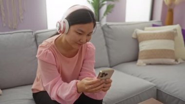 Oturma odasındaki kanepede akıllı telefon kullanan, gülümseyen, rahatlatan ve evde müzik dinlemekten zevk alan bir kadın..