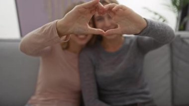 Sıcak oturma odasında oturup aile sevgisini ve birlikteliğini aktaran kız kardeşler elleriyle kalp şekli oluşturuyorlar.