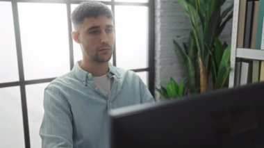 Genç adam, bilgisayar ekranına odaklanırken bitkiler ve büyük pencerelerle modern ofiste endişeli bir şekilde oturuyor..