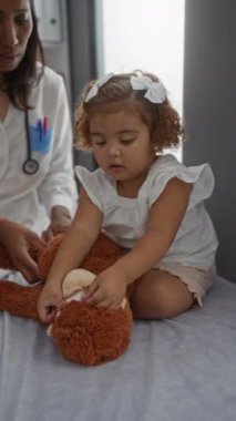 Klinikte kız arkadaşı olan bir kadın çocuk doktoru Teddy ile oynuyor, kapalı bir tıbbi ortamda çocuk sağlığını resmediyor, doktor-hasta etkileşimini vurguluyor..