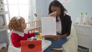 Anne belgelerini okurken, kız evde tablet izliyor, oyuncaklarla çevrili, rahat ve sakin bir yatak odası ortamında, modern aile hayatını temsil ediyor..
