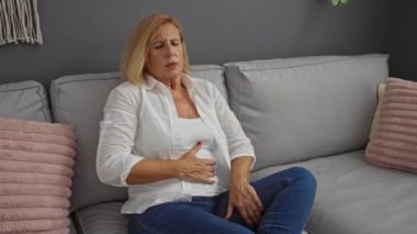 Oturma odasındaki koltukta otururken karın ağrısı çeken bir kadın, gri bir arka planda, olgun sarışın bir kadınla ev ortamını düzenliyor..