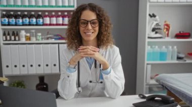 Klinikte gülümseyen, gözlüklü, laboratuvar önlüklü, kıvırcık saçlı, tıbbi ortamında ve profesyonel atmosferde steteskop kullanan bir kadın..