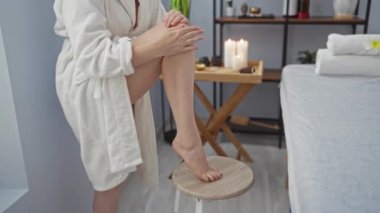 Kadın sağlık merkezine losyon sürüyor, beyaz bir bornoz giyiyor, yanan bir mum eşliğinde.