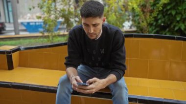 Şehir parkında sarı bir bankta oturan genç adam, akıllı telefonuna odaklanmış, gündüzleri şehir merkezinde günlük kıyafetler giyiyor..