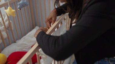 Kadın bebek beşiğini rahat yatak odasına yerleştiriyor. Huzurlu bir ev ortamında aile bakımını ve sevgiyi ön plana çıkarıyor..