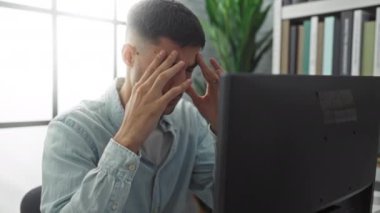 Ofiste stresli görünen genç bir adam, bilgisayarlı bir masada oturuyor, elleri şakakların üzerinde, kitaplar ve bitkilerle çevrili, büyük pencerelerden gelen doğal ışıkla aydınlatılmış..