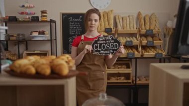 Etrafında çeşitli ekmek ve hamur işleri olan bir fırında açık tabela tutan kadın.