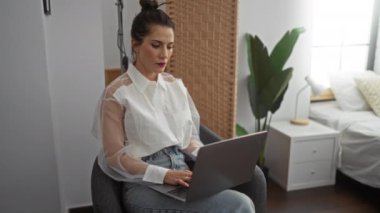 Yatak odasında oturan bir kadın dizüstü bilgisayar kullanırken endişeli görünüyor, eli alnında endişeli bir ifade sergiliyor..