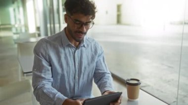 Modern ofis ortamında gözlüklü genç adam, tablet kullanıyor, çizgili gömlek giyiyor, elinde kahve tutuyor, gülümsüyor, doğal ışık, cihaza odaklanıyor, günlük iş ayarlarına odaklanıyor..