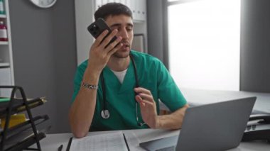 Steteskoplu önlüklü genç adam hastane kliniğinde akıllı telefon ve dizüstü bilgisayar kullanıyor..
