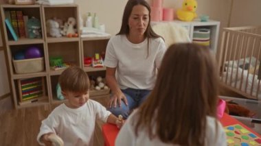 İki çocukla rahat bir yatak odasında oyuncaklarla oynayan, ev ortamında aile etkileşimlerini sergileyen, kapalı mekanda oyun oynayarak ve atmosferi geliştirerek vurgulayan bir kadın..