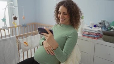 Yatak odasında oturan hamile bir kadın akıllı telefona gülümsüyor. Etrafı yumuşak bebek kıyafetleri ve rahat dekorlarla çevrili bir rahatlama anının tadını çıkarıyor..