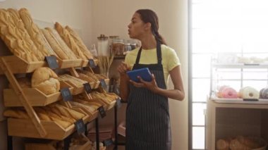 Bir pastanede elinde tablet tutan genç bir kadın. Kapalı alandaki raflarda ekmek sergiliyor.