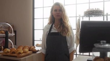 Pastanede önlük giymiş sarışın bir kadın tezgahın arkasında elinde hamur işleriyle dikilirken başparmağını kaldırıyor.