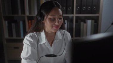 Ofiste çalışan bir kadın, arka planında kitaplık bulunan bir kulaklık kullanarak profesyonel bir iş ortamı ve video iletişimi kuruyor.