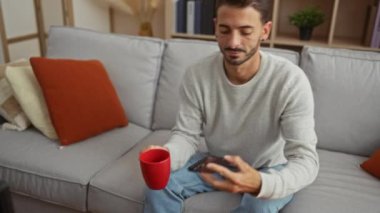 Genç adam evinde rahat bir kanepede kırmızı bir kupa tutuyor ve akıllı telefon kullanıyor. Etrafı renkli yastıklarla ve oturma odasındaki raflarla çevrili..